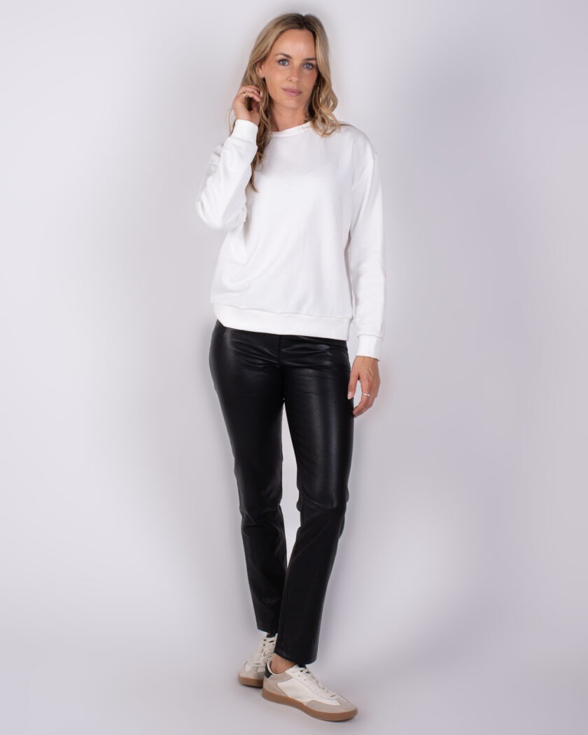 Vrouw in witte trui, zwarte leren broek en sneakers poseert voor een neutrale achtergrond.