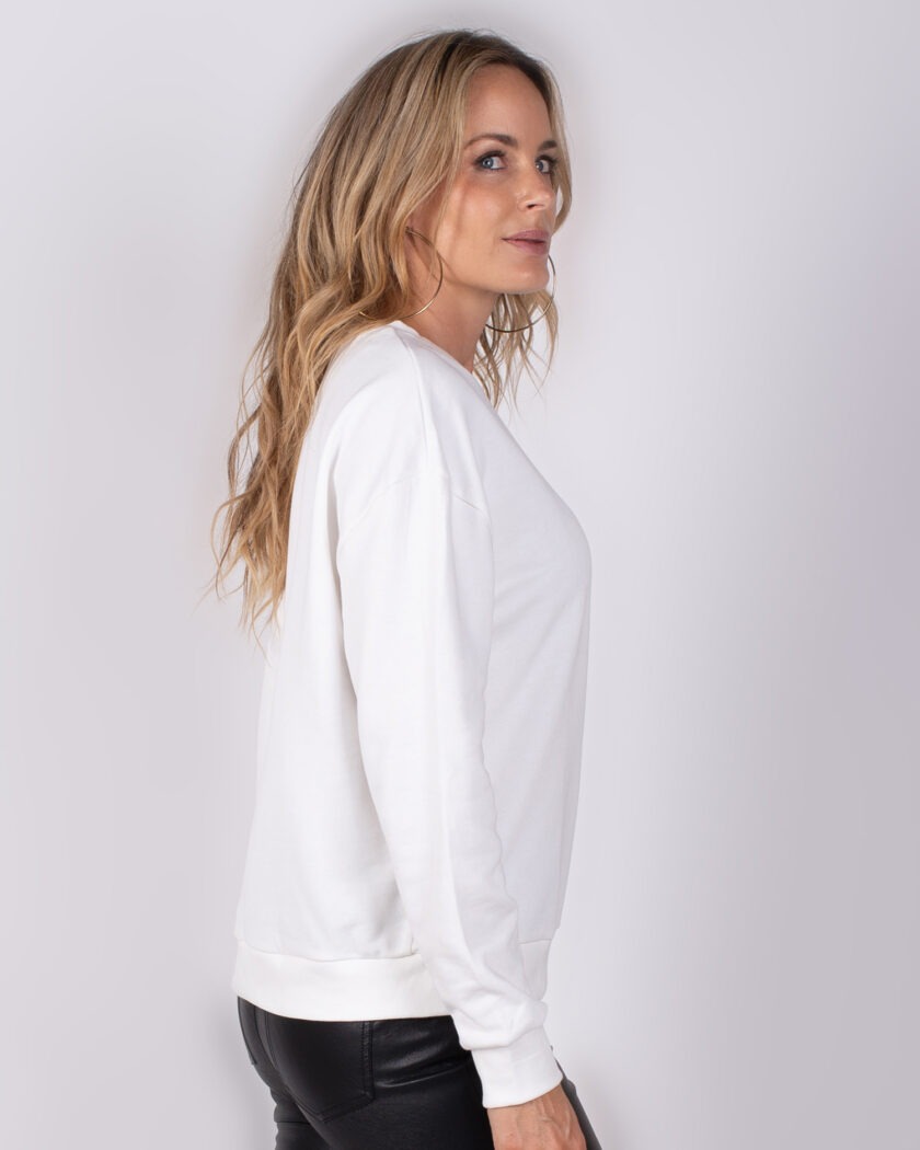 Vrouw in zijaanzicht draagt een witte trui uit de Studio AW25-26 collectie en zwarte leren broek.