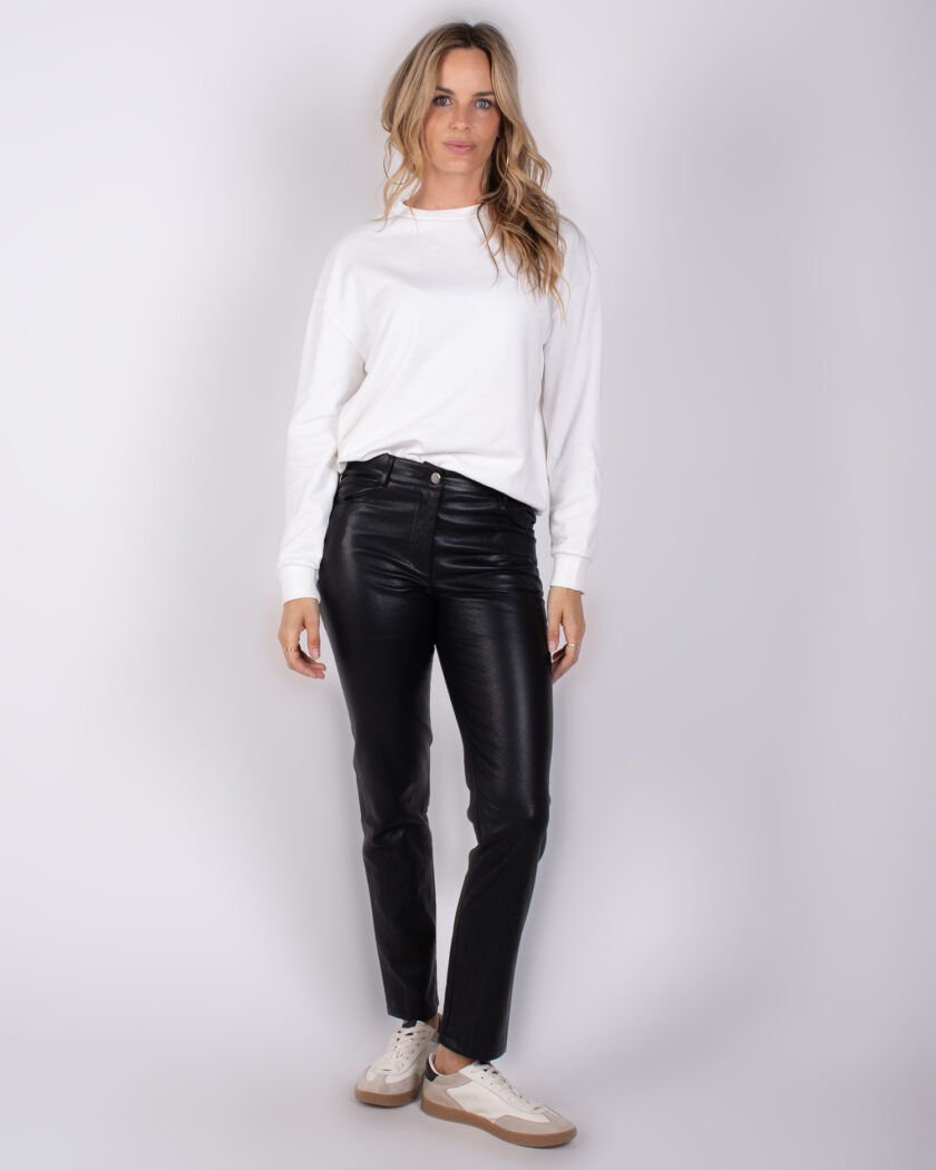 Vrouw in een witte trui en zwarte leren broek uit Studio_AW25-26#3-61 collectie, poserend tegen een neutrale achtergrond.