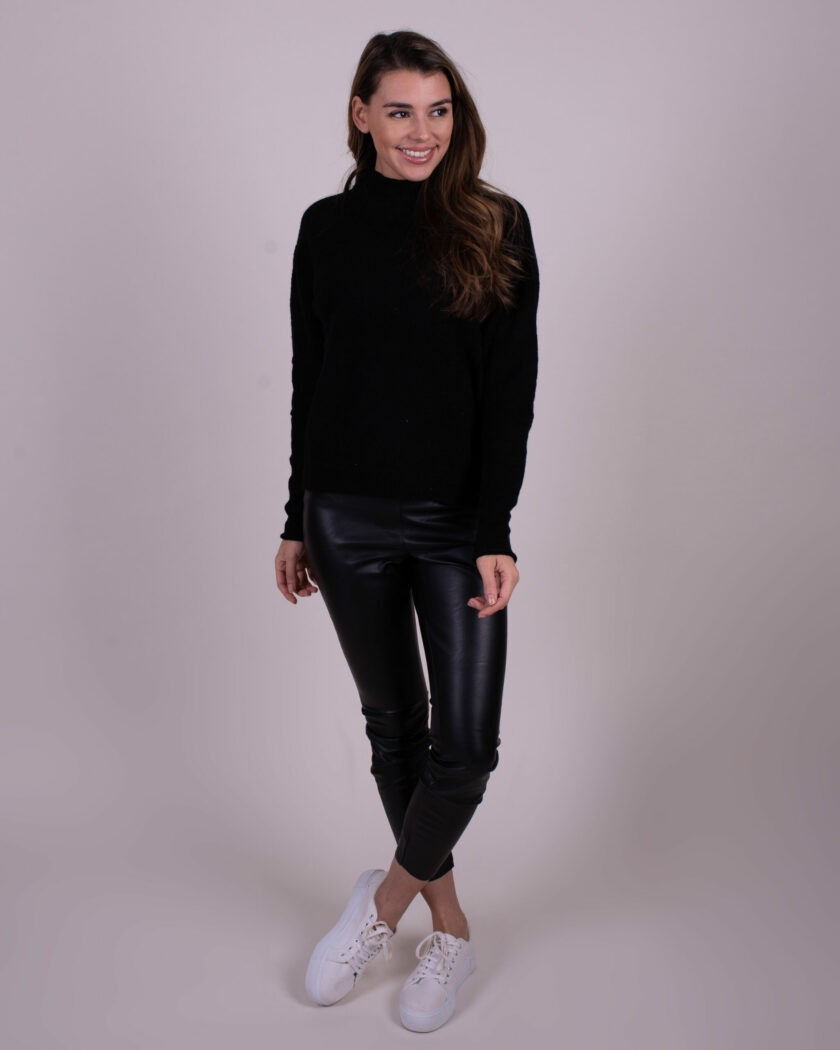 Vrouw in zwarte trui en zwarte leren broek poseert met witte sneakers tegen een neutrale achtergrond.