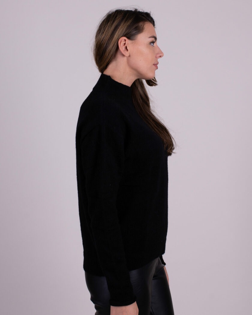 Vrouw in zijaanzicht draagt een zwarte trui en zwarte leren broek tegen een neutrale achtergrond.