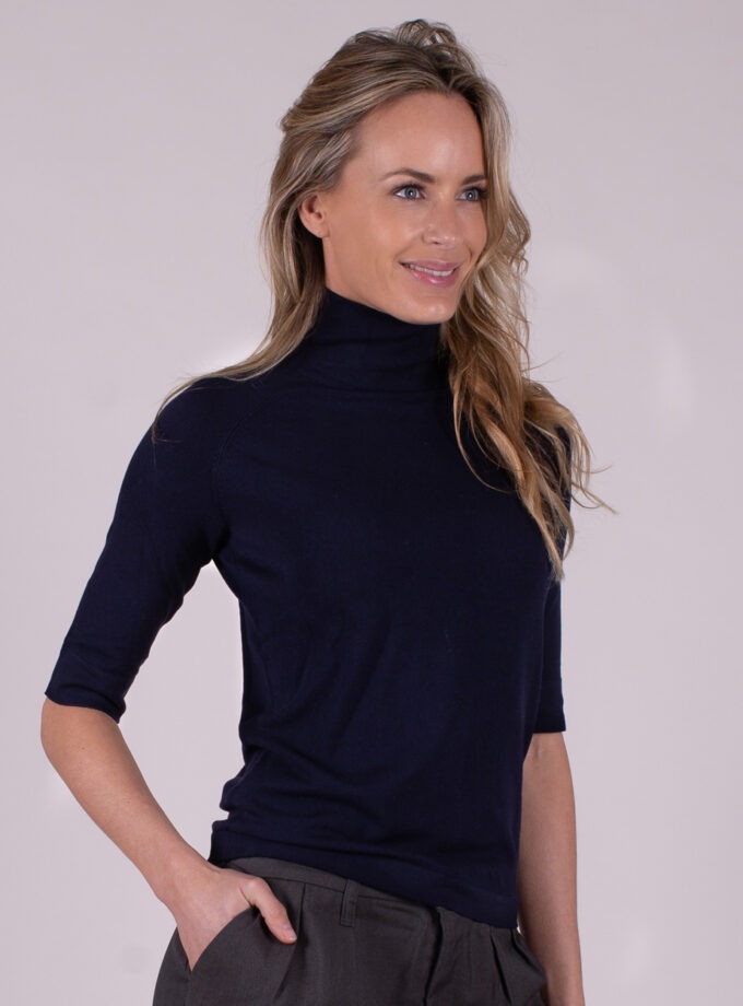 Vrouw draagt een donkerblauwe coltrui met halve mouwen en grijze broek in een studiosetting.