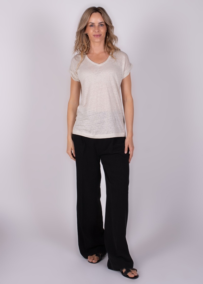Een vrouw draagt een lichtbeige T-shirt en zwarte wijde broek tegen een neutrale achtergrond.