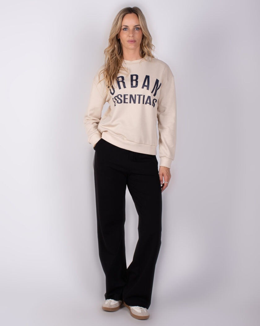 Vrouw in casual outfit met beige sweater met de tekst "Urban Essentials", zwarte broek en sneakers.
