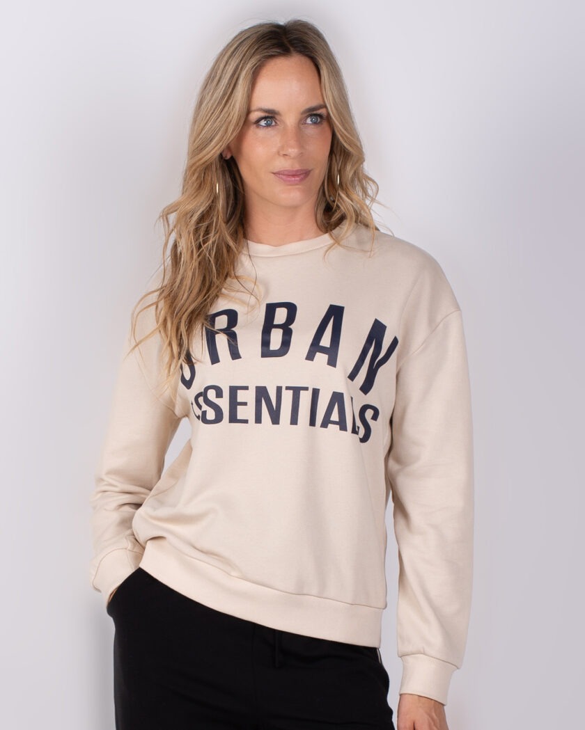 Vrouw draagt een beige sweatshirt met de tekst "URBAN ESSENTIALS" en zwarte broek, tegen een neutrale achtergrond.