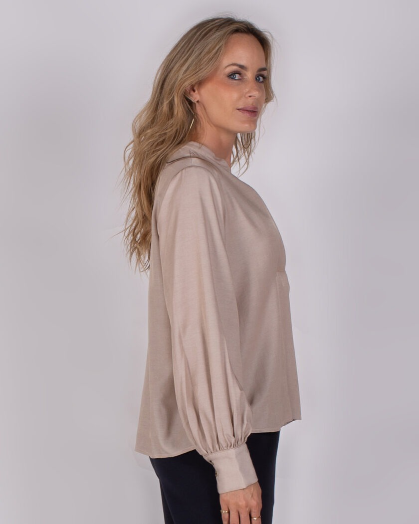 Vrouw draagt een beige blouse met pofmouwen uit de Studio AW25-26 collectie, gefotografeerd van opzij tegen een neutrale achtergrond.