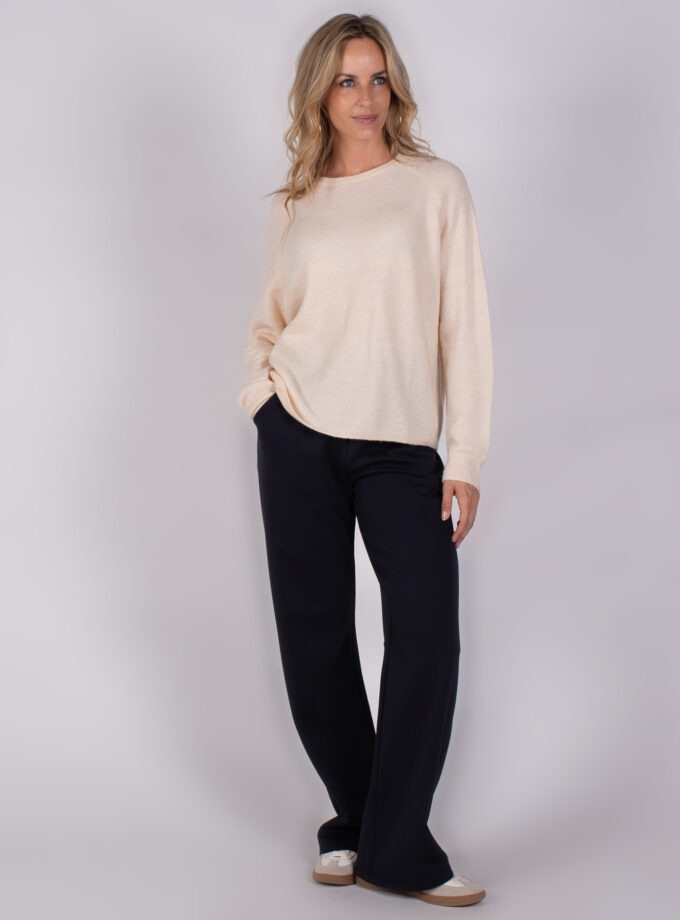 Vrouw met los blond haar draagt een beige trui en zwarte wijde broek uit de Studio AW25-26 collectie.