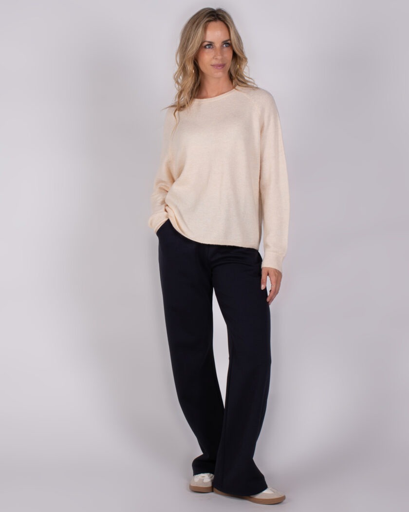 Vrouw met los blond haar draagt een beige trui en zwarte wijde broek uit de Studio AW25-26 collectie.