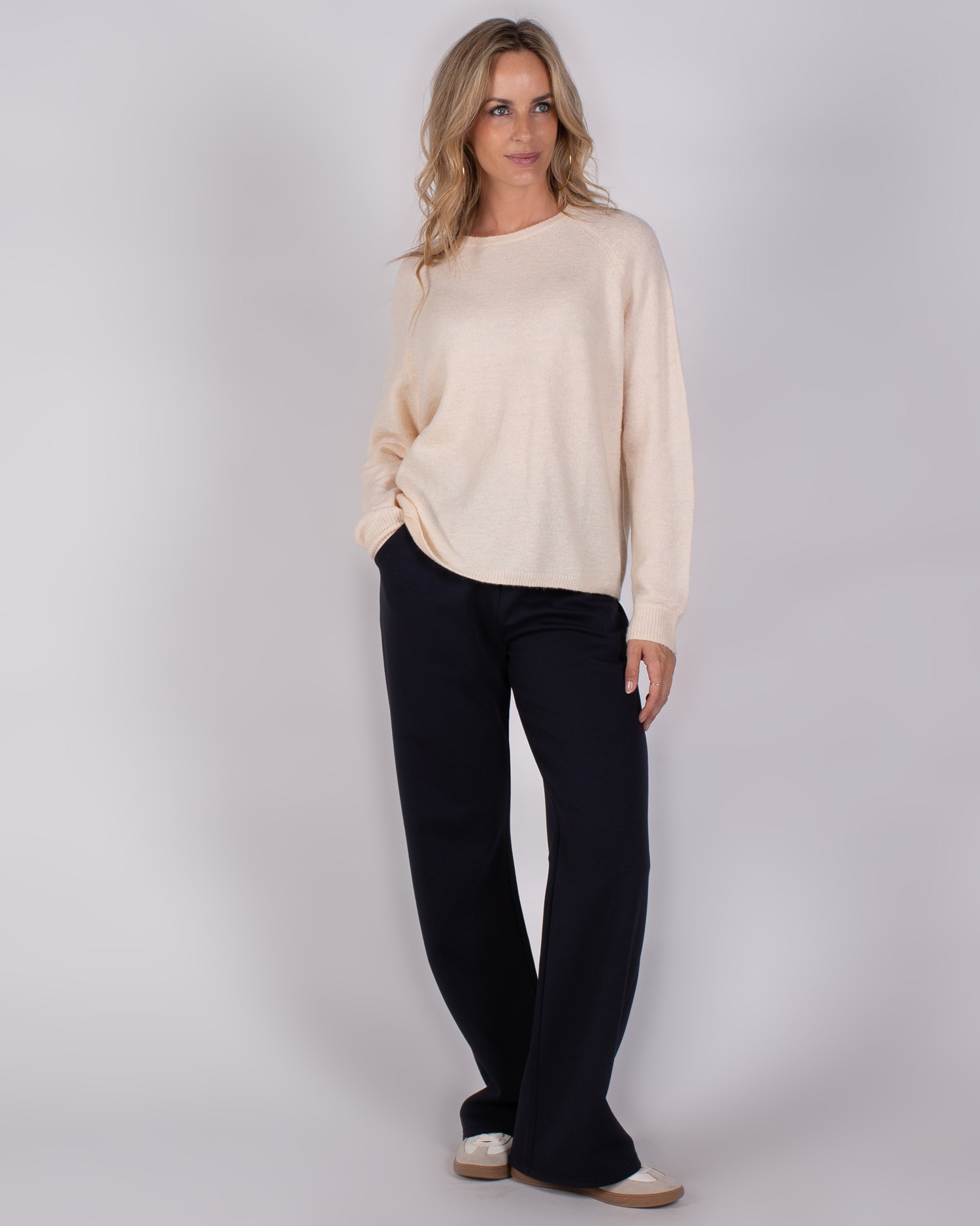 Vrouw met los blond haar draagt een beige trui en zwarte wijde broek uit de Studio AW25-26 collectie.