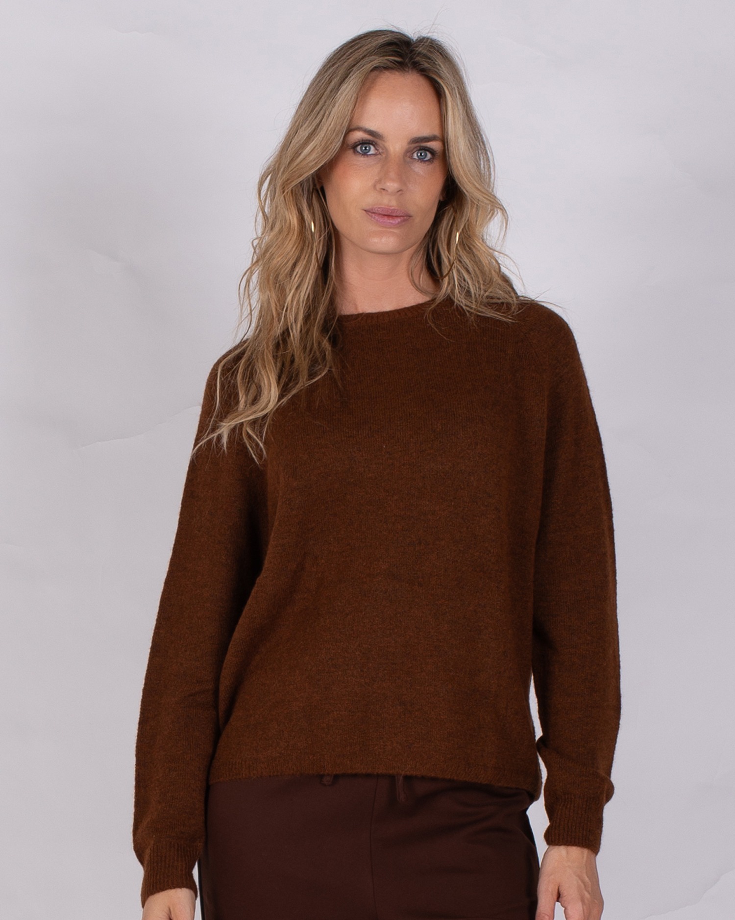 Vrouw draagt een donkerbruine trui uit de Studio AW25-26 collectie tegen een neutrale achtergrond.