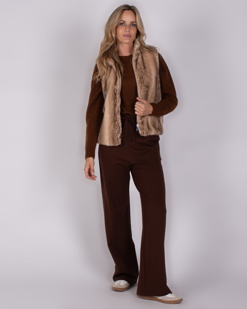 Vrouw draagt een winteroutfit met een bruine trui, wijde donkerbruine broek en een beige faux fur bodywarmer uit de Studio AW25-26 collectie.