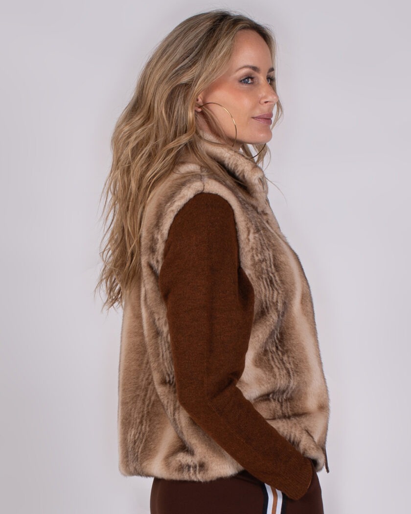 Vrouw in profiel met lang blond haar draagt een bruin Studio AW25-26-124 vest van imitatiebont over een bruine trui.