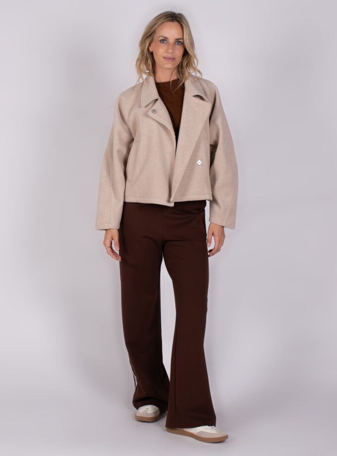 Vrouw draagt een beige jas en bruine broek uit de Studio AW25-26 collectie tegen een neutrale achtergrond.