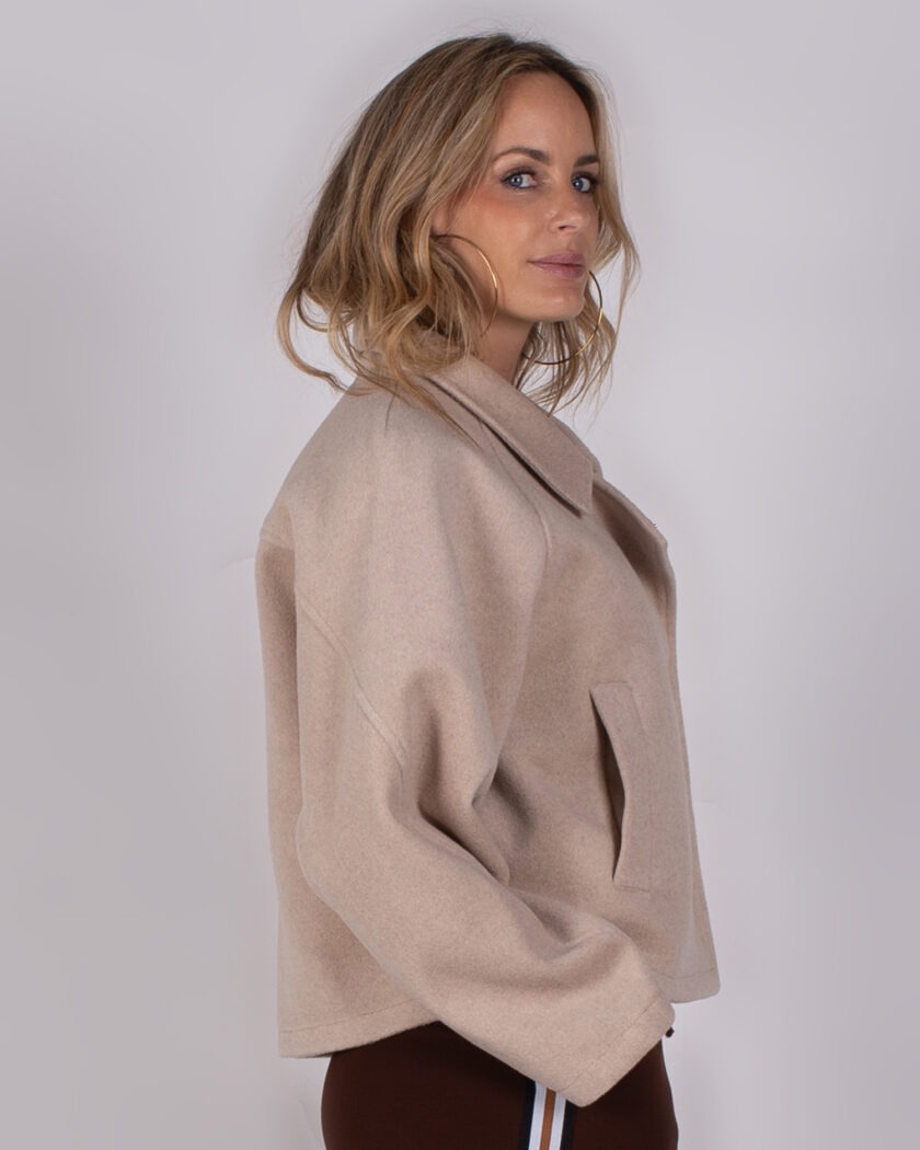 Vrouw in beige, wollen jas uit de Studio AW25-26 collectie poseert zijwaarts tegen een neutrale achtergrond.