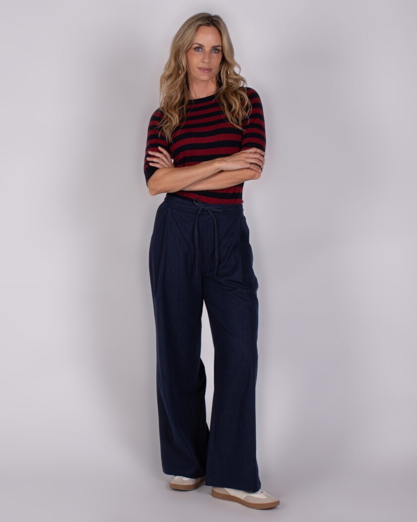 Vrouw in donkerblauw-rood gestreepte top en wijde donkerblauwe broek poseert met gekruiste armen tegen een neutrale achtergrond.