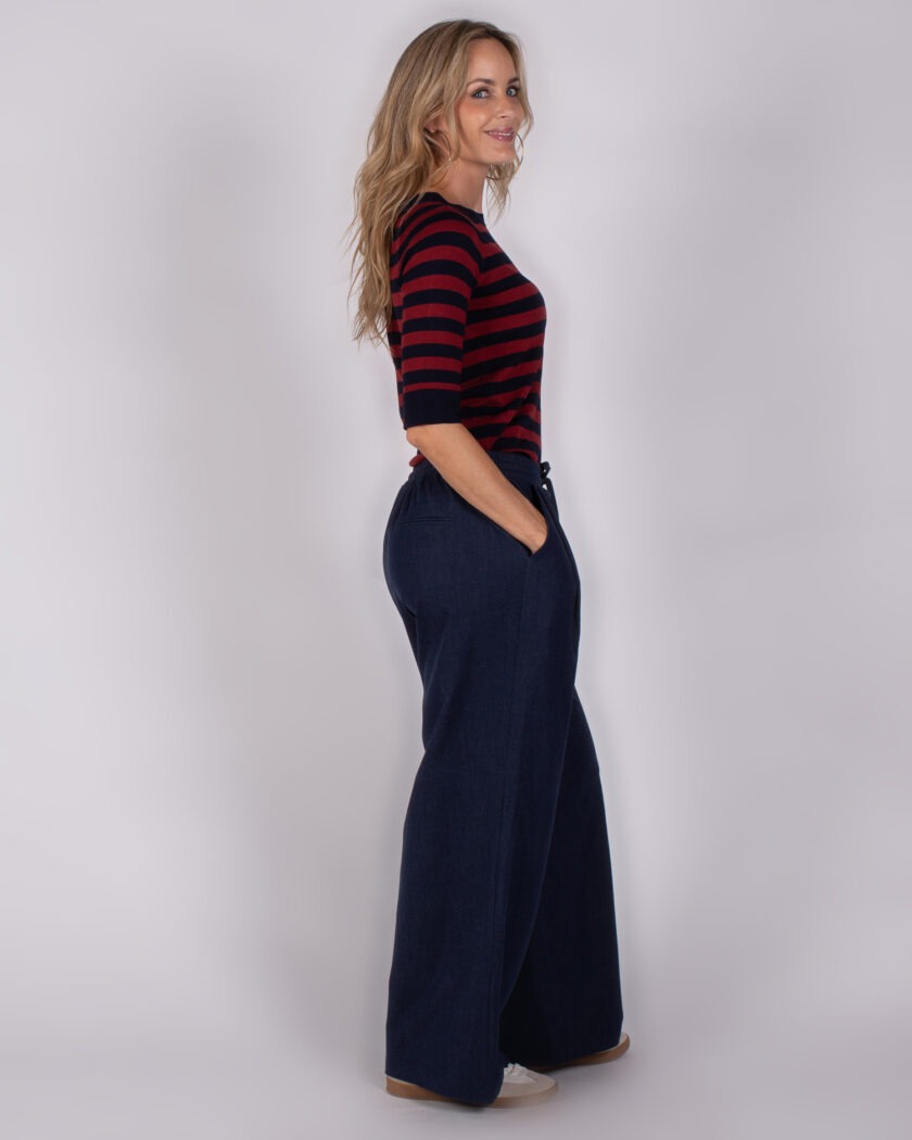 Vrouw in zijaanzicht draagt een rood-zwart gestreepte top en wijde donkerblauwe broek met handen in de zakken.