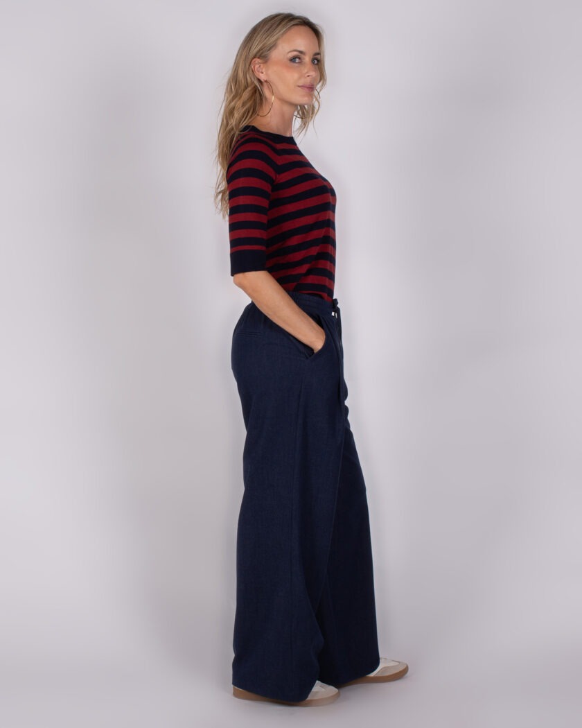 Vrouw in zijaanzicht draagt een rood-blauw gestreepte top en wijde donkerblauwe broek uit de Studio AW25-26 collectie.