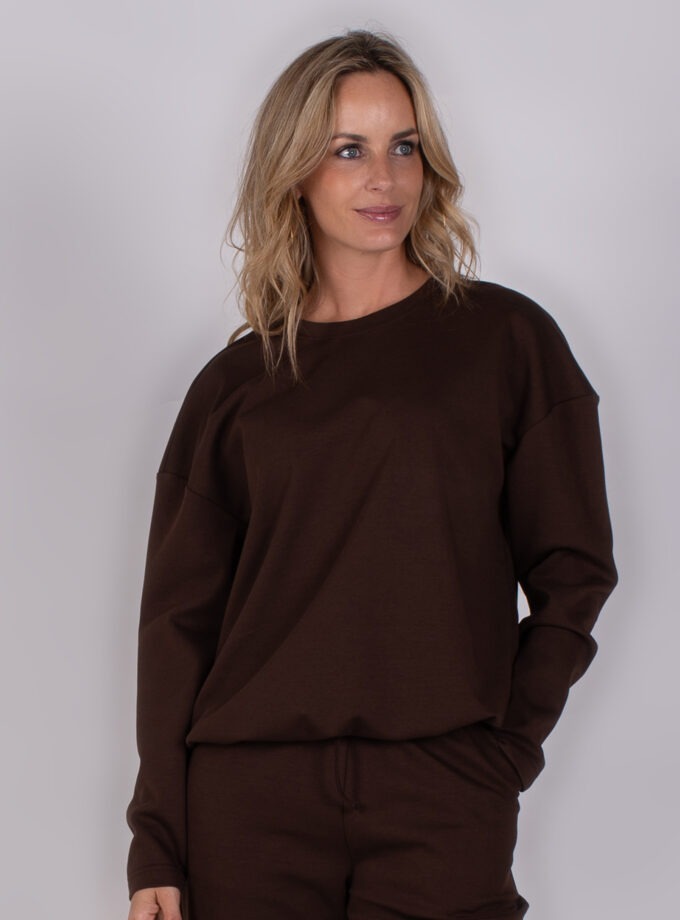 Vrouw draagt een donkerbruin loungeset met lange mouwen uit de Studio AW25-26 collectie.