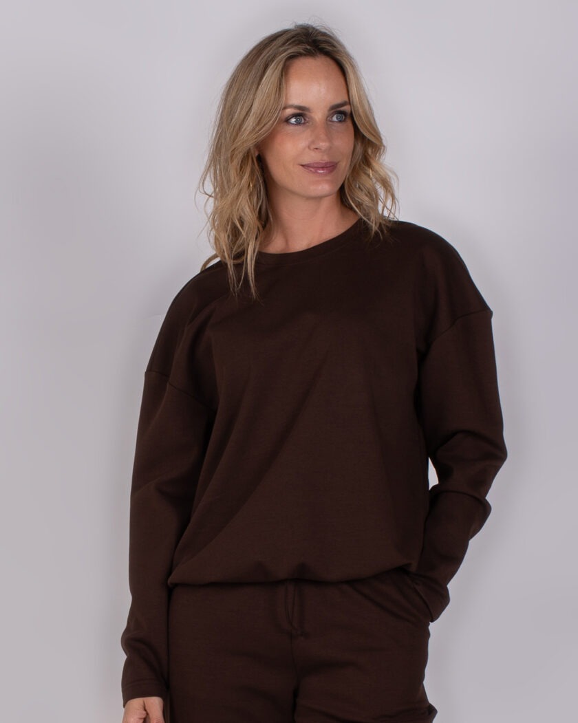 Vrouw draagt een donkerbruin loungeset met lange mouwen uit de Studio AW25-26 collectie.
