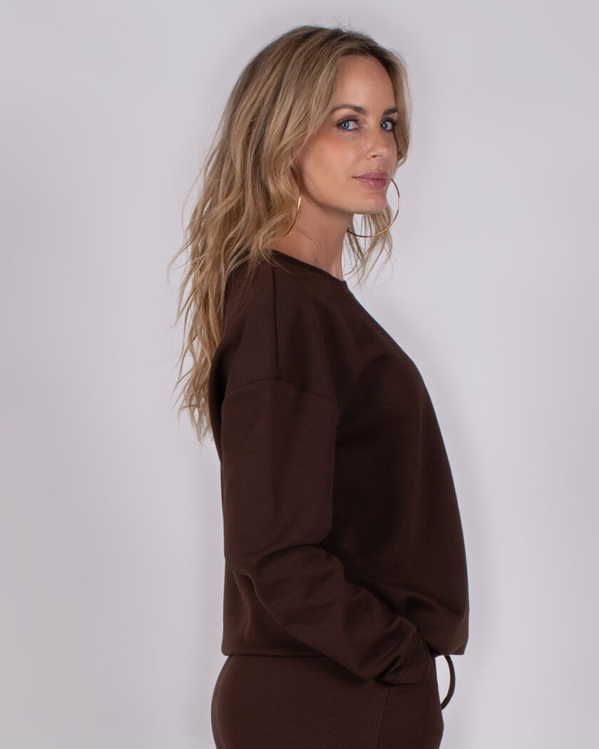 Vrouw in zijaanzicht draagt een donkerbruine sweater en bijpassende broek uit Studio AW25-26 collectie.