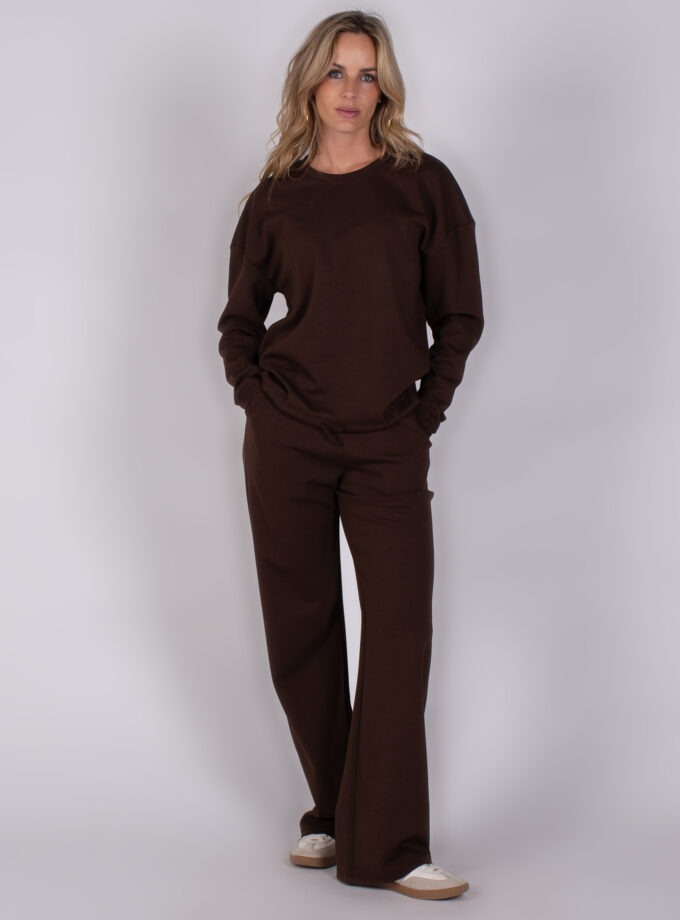Vrouw draagt een donkerbruin loungepak met wijde broek en sneakers uit de Studio AW25-26 collectie.
