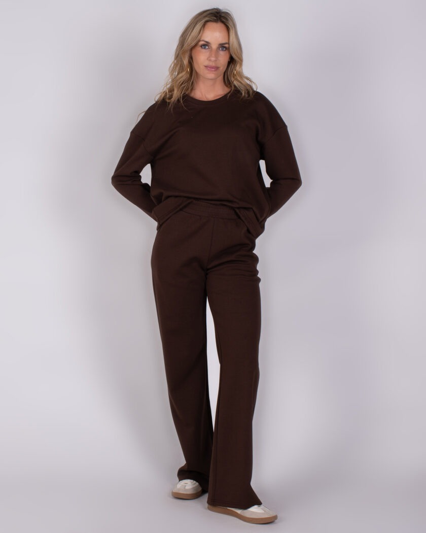 Vrouw draagt een donkerbruin loungpak bestaande uit een trui met lange mouwen en een wijde broek, uit de Studio AW25-26 collectie.