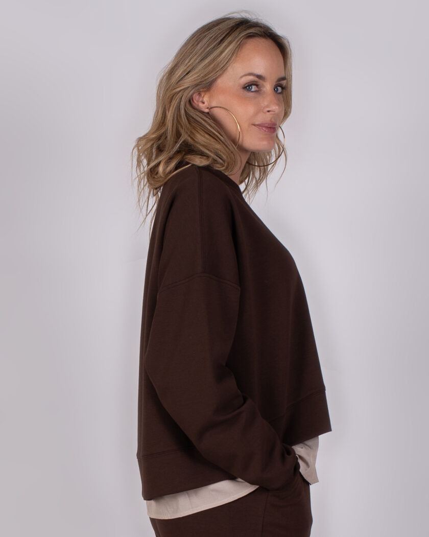 Vrouw in een donkerbruine oversized trui uit de Studio AW25-26 collectie kijkt met een glimlach over haar schouder.