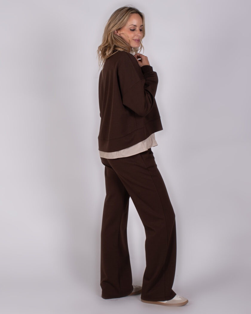 Vrouw poseert in een comfortabele, donkerbruine loungewear-set uit de Studio AW25-26 collectie tegen een neutrale achtergrond.