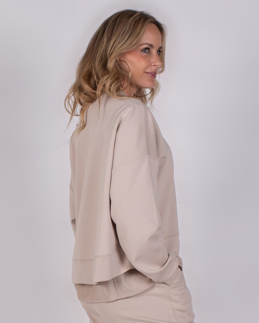 Vrouw met halflang blond haar draagt een beige oversized loungepak uit de Studio AW25-26-191 collectie.