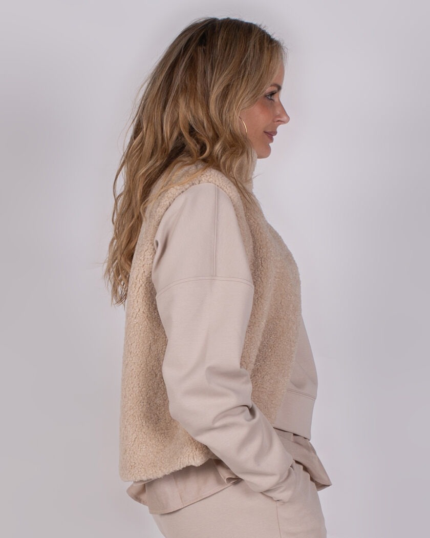 Vrouw in beige outfit met een teddy bodywarmer, gefotografeerd van opzij tegen een neutrale achtergrond.