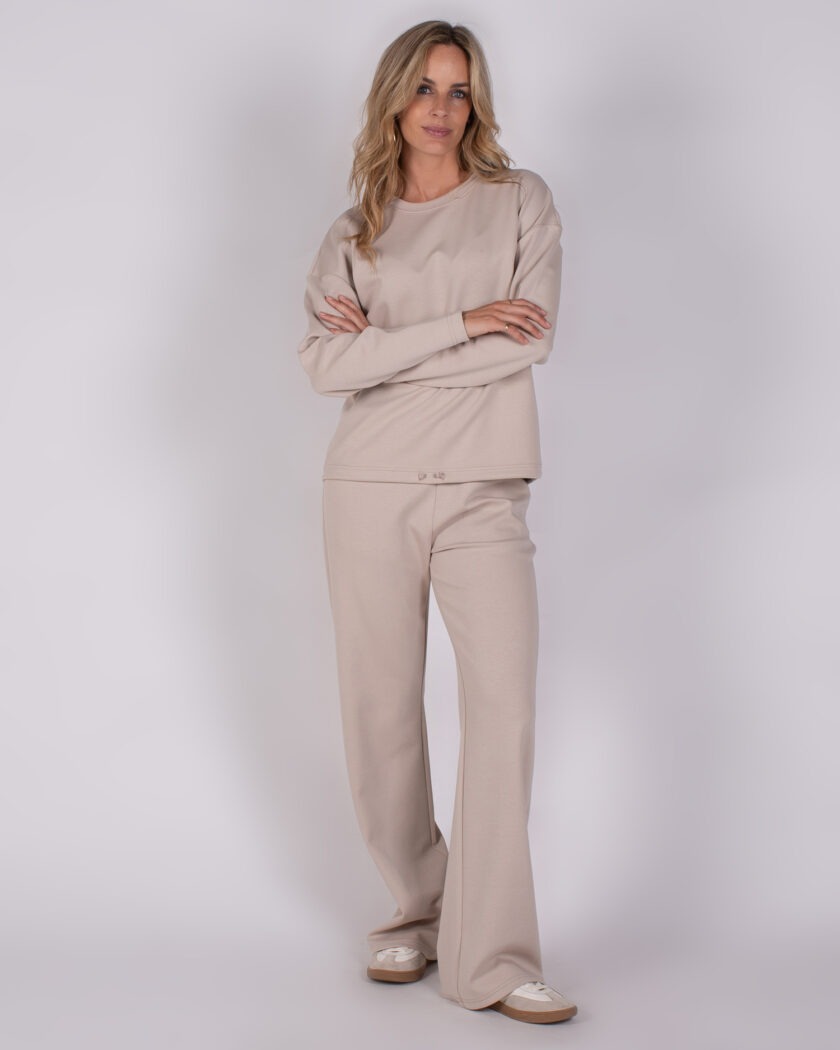 Vrouw draagt een beige loungewear-set bestaande uit een trui met lange mouwen en wijde broek uit de Studio AW25-26 collectie.