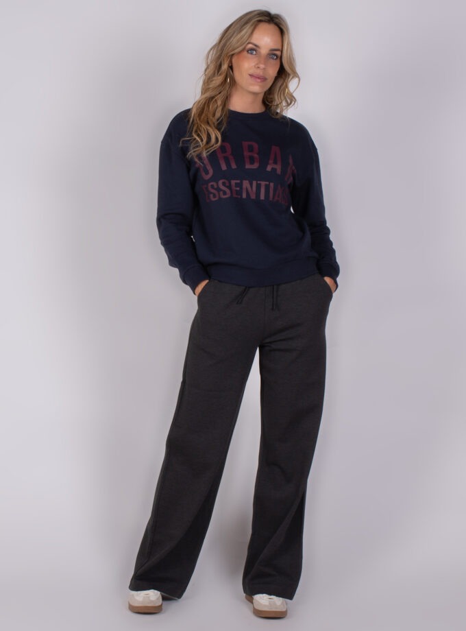 Vrouw draagt een donkerblauwe trui met de tekst "Urban Essentials" en losse, donkere loungebroek uit de Studio AW25-26 collectie.