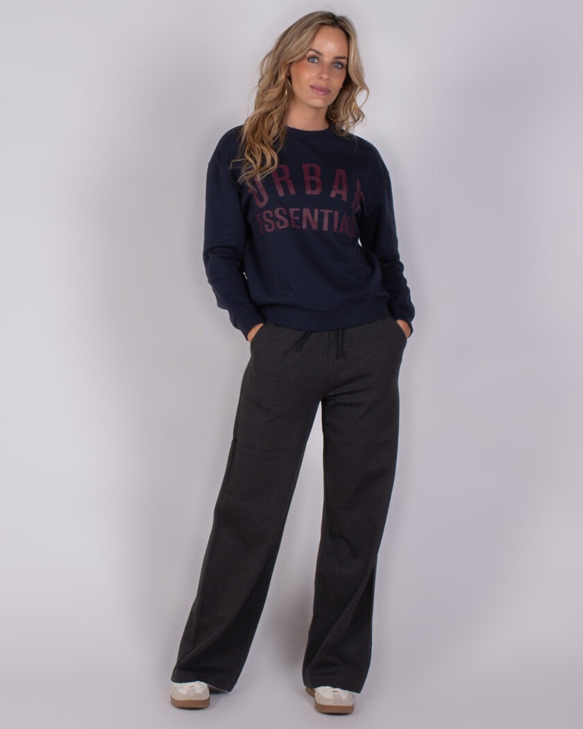 Vrouw draagt een donkerblauwe trui met de tekst "Urban Essentials" en losse, donkere loungebroek uit de Studio AW25-26 collectie.