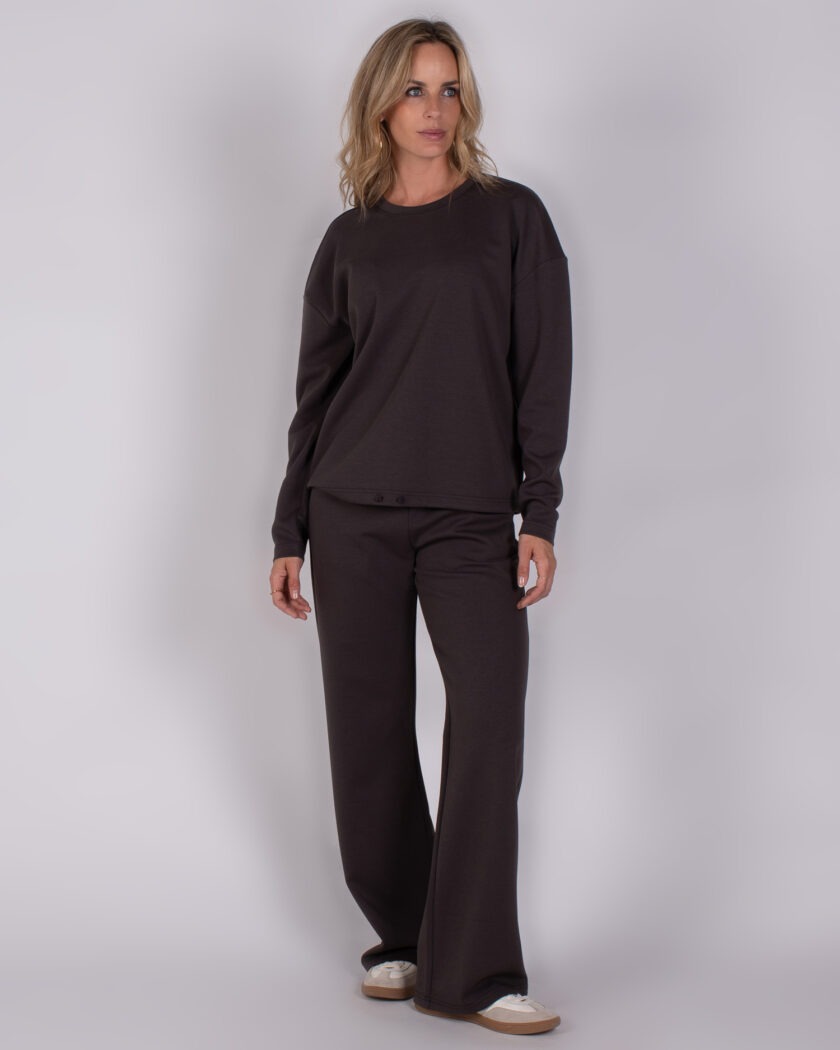 Vrouw draagt een donkerbruin loungpak met lange mouwen en wijde broek uit de Studio AW25-26 collectie.