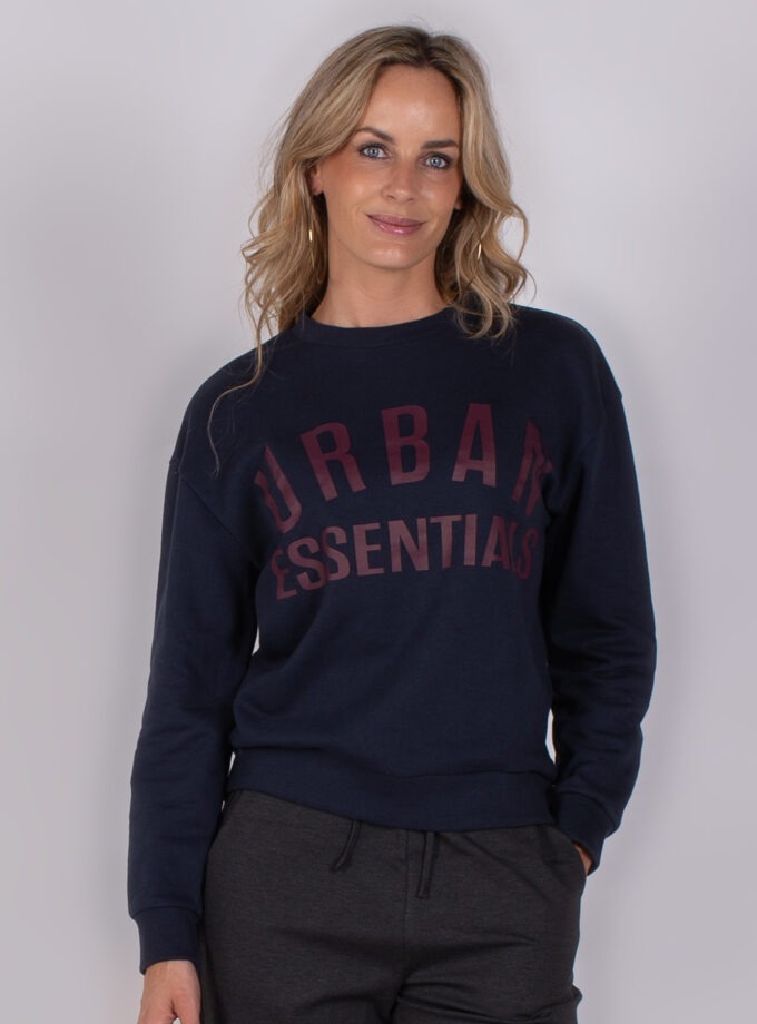 Vrouw draagt een donkerblauwe sweater met de tekst "Urban Essentials" uit de Studio AW25-26-22 collectie.