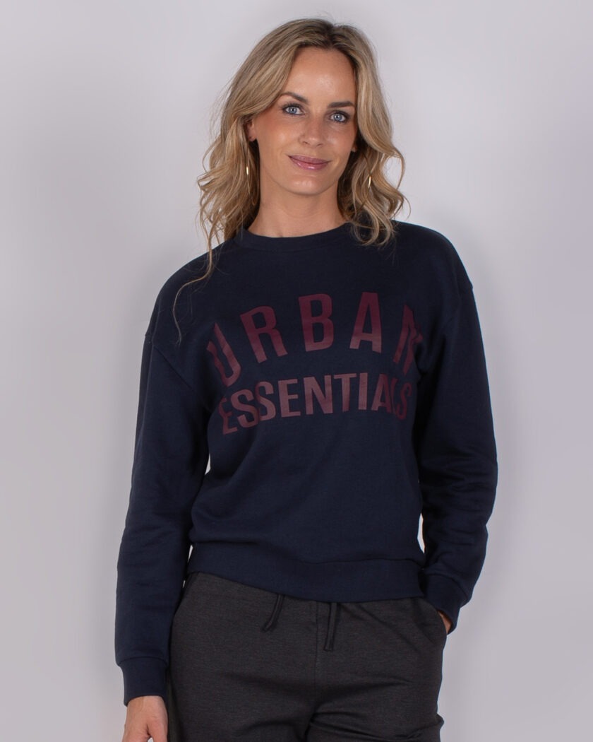 Vrouw draagt een donkerblauwe sweater met de tekst "Urban Essentials" uit de Studio AW25-26-22 collectie.