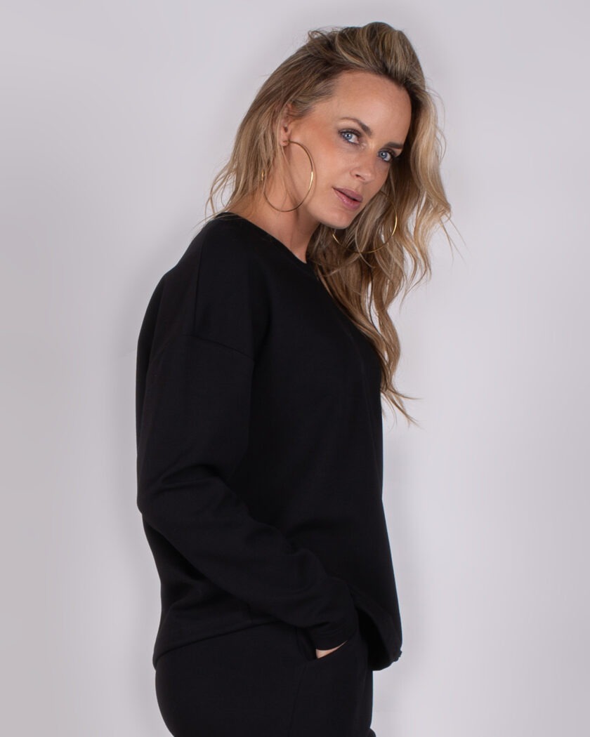 Vrouw in zwarte Studio_AW25-26-222 trui en broek poseert zijwaarts tegen een neutrale achtergrond.