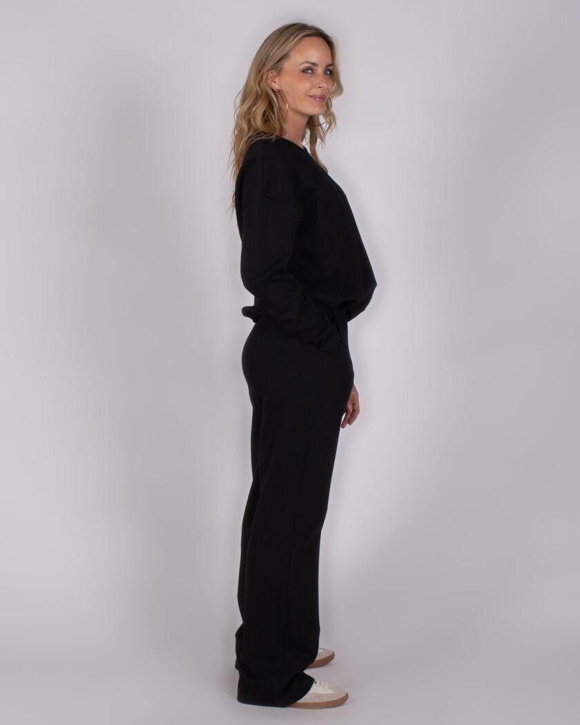 Vrouw in zijaanzicht draagt een zwarte Studio_AW25-26-230 set met wijde broek en trui.