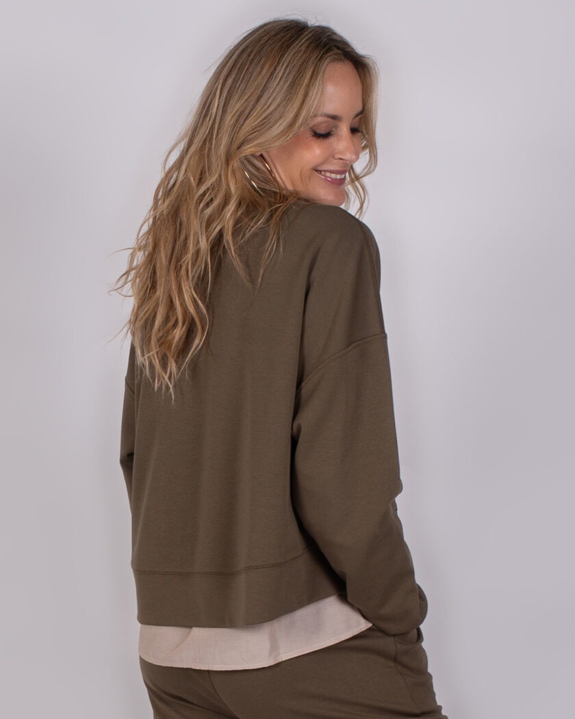 Vrouw met lang blond haar draagt een olijfgroene trui en broek uit de Studio AW25-26 collectie.