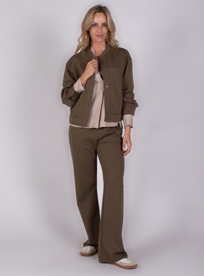 Vrouw draagt een bruin Studio_AW25-26-239 broekpak met beige blouse en sneakers tegen een neutrale achtergrond.