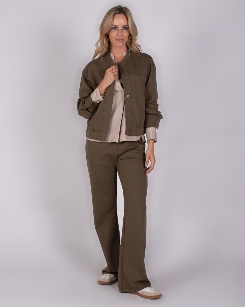 Vrouw draagt een bruin Studio_AW25-26-239 broekpak met beige blouse en sneakers tegen een neutrale achtergrond.