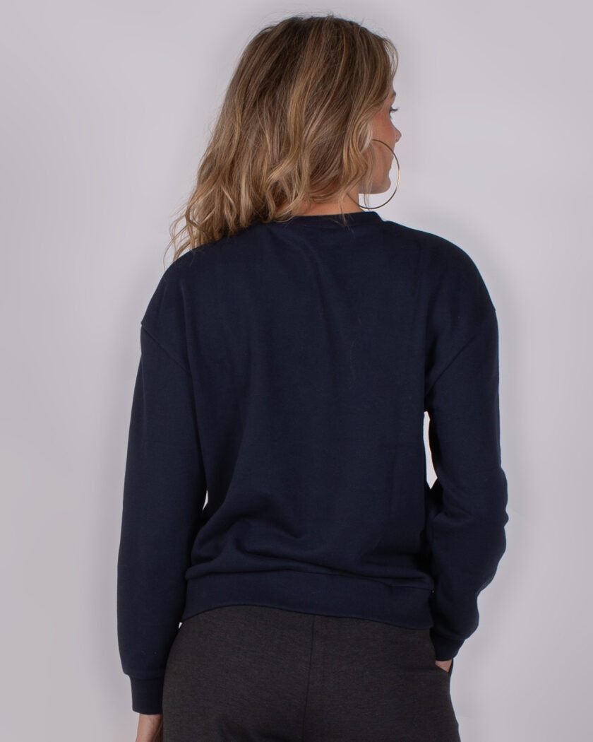 Vrouw met lang blond haar draagt een donkerblauwe trui en donkere broek, gefotografeerd van achteren tegen een neutrale achtergrond.
