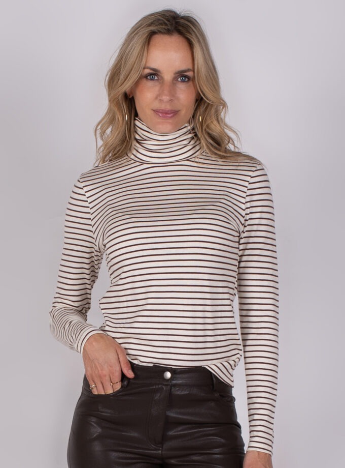 Vrouw met halflang blond haar draagt een beige coltrui met zwarte strepen en een zwarte leren broek.