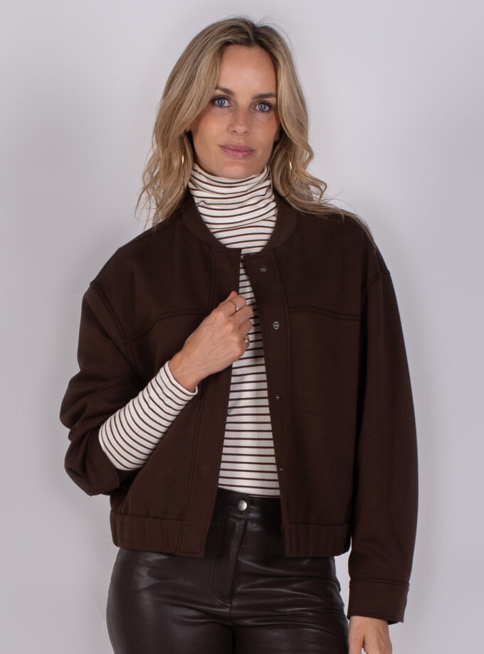Vrouw met halflang blond haar draagt een donkerbruine jas over een gestreepte coltrui en een zwarte leren broek.