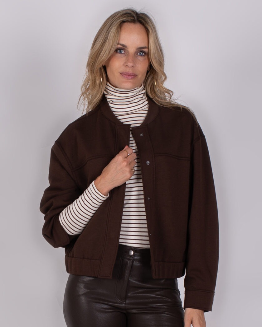 Vrouw met halflang blond haar draagt een donkerbruine jas over een gestreepte coltrui en een zwarte leren broek.