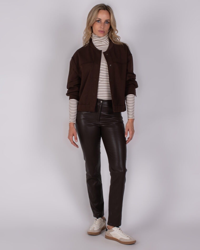 Vrouw draagt een donkerbruine jas met bijpassende leren broek, gestreepte coltrui en witte sneakers uit de Studio AW25-26 collectie.