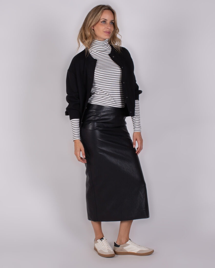 Vrouw draagt een zwart-witte gestreepte coltrui, zwarte midi-rok van leer, zwart jasje en witte sneakers.