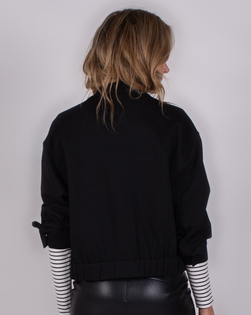 Achteraanzicht van een vrouw in een zwarte Studio AW25-26-302 jas met opgerolde mouwen en een gestreepte top eronder.