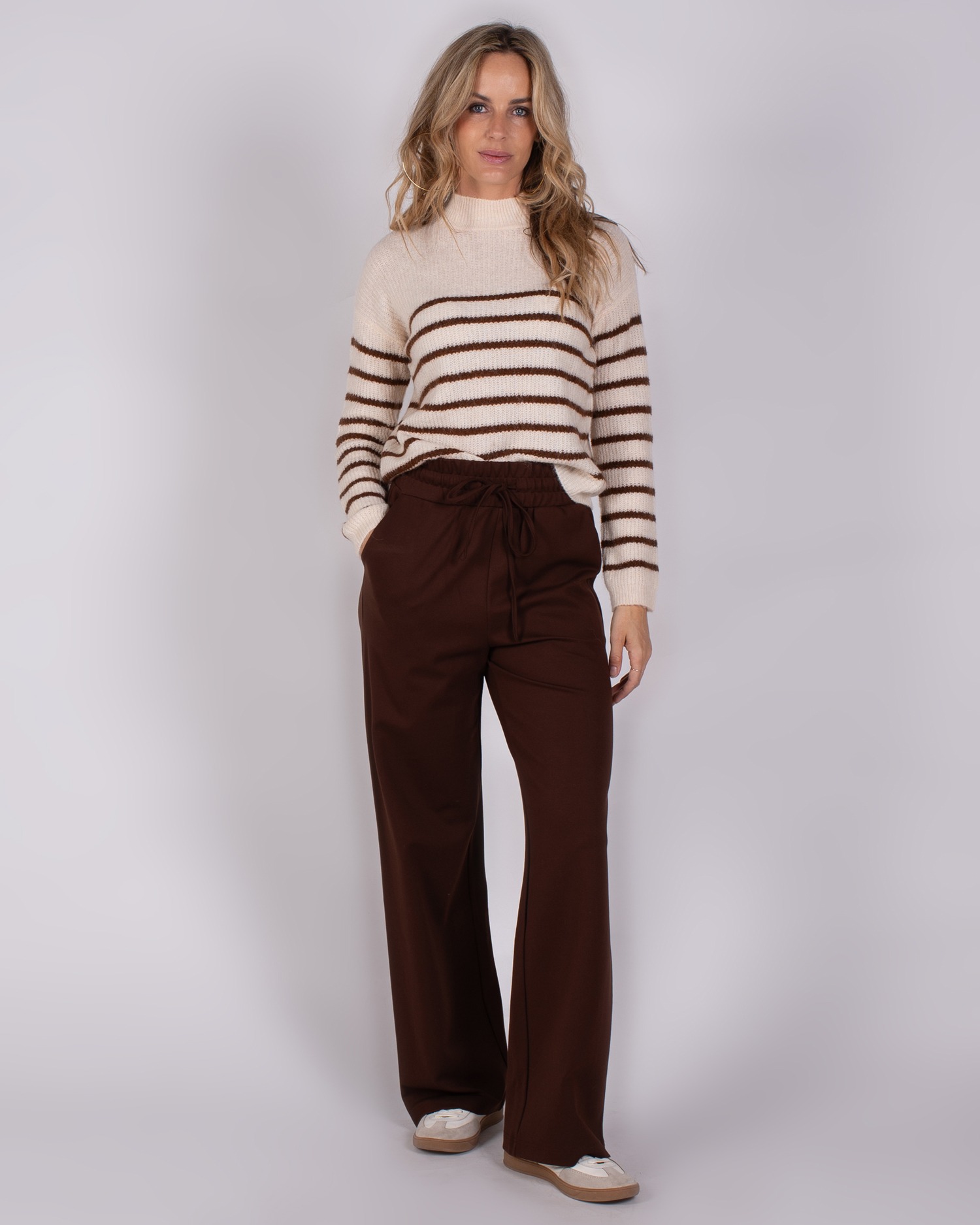 Vrouw draagt een crèmekleurige trui met bruine strepen en wijde, donkerbruine broek uit de Studio AW25-26 collectie.