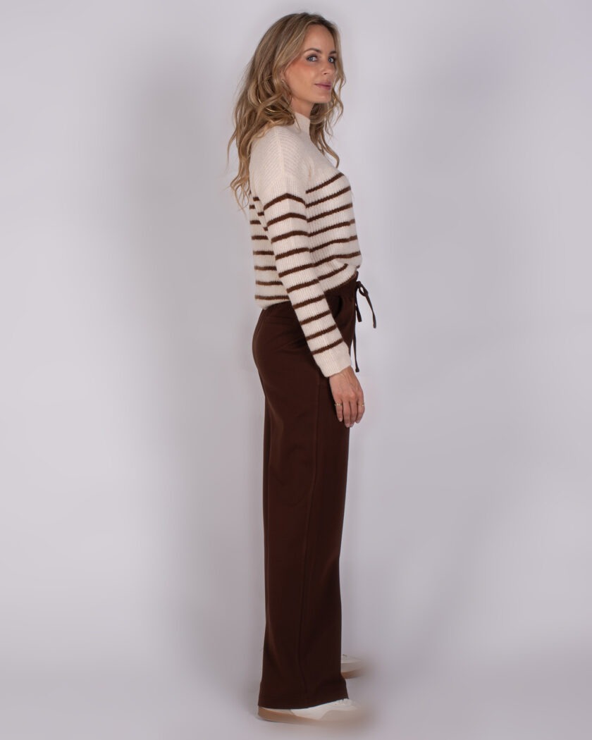 Vrouw in zijaanzicht draagt een wit-beige trui met bruine strepen en bruine broek uit de Studio AW25-26 collectie.